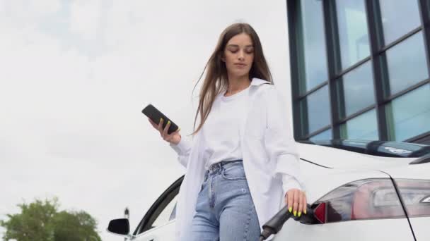 Young Woman Standing Electric Car Looks Smart Phone — Vídeo de stock