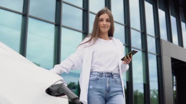 若い女性が電気自動車の近くに立っていて スマートフォンを見ている レンタカーは電気自動車の充電ステーションで充電しています 車の共有 — ストック動画