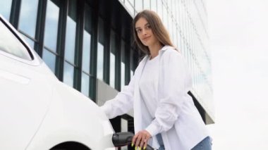 Güneşli alışveriş merkezinin otoparkında elektrik kablosu takan gülümseyen beyaz kız, seçici odaklanma. Yaşam tarzı ve ekoloji kavramı