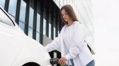 Güneşli alışveriş merkezinin otoparkında elektrik kablosu takan gülümseyen beyaz kız, seçici odaklanma. Yaşam tarzı ve ekoloji kavramı