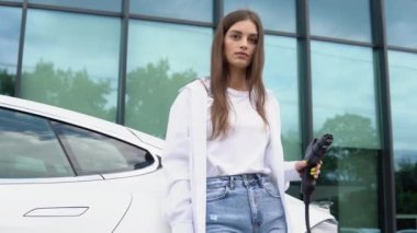 Güneşli alışveriş merkezinin otoparkında elektrik kablosu takan gülümseyen beyaz kız, seçici odaklanma. Yaşam tarzı ve ekoloji kavramı.