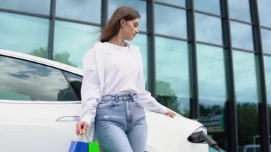 Happy young girl standing on city parking near electric car, charging automobile battery from small city station, holding shopping bags and watching smart phone