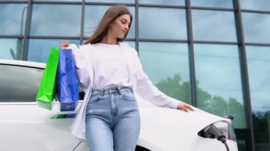 Young girl after buying products with a shopping bag is standing near the charging electric car