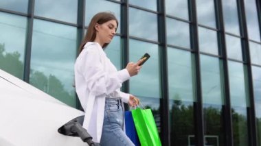 Happy young girl standing on city parking near electric car, charging automobile battery from small city station, holding shopping bags and watching smart phone