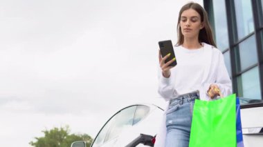 Young girl after buying products with a shopping bag is standing near the charging electric car