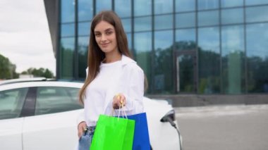 Young girl after buying products with a shopping bag is standing near the charging electric car