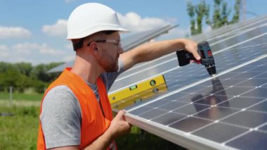 Male engineer in protective helmet installing solar photovoltaic panel system using screwdriver. Electrician mounting blue solar module on roof of modern house