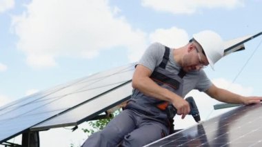 Male engineer in protective helmet installing solar photovoltaic panel system using screwdriver. Electrician mounting blue solar module on roof of modern house. Alternative energy ecological concept