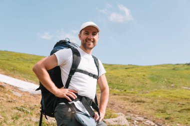 A bearded traveler with a backpack on the top of a mountain. A tourist with a backpack stands against the background of a mountain.