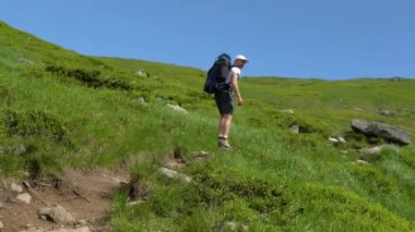 Male hiker backpacker with trekking poles reaches spot overlooking beautiful mountains. Man traveler standing on rock looking at incredible highland landscape on sunny day. Mountain peaks