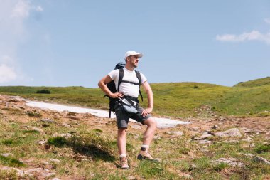 A bearded traveler with a backpack on the top of a mountain. A tourist with a backpack stands against the background of a mountain.