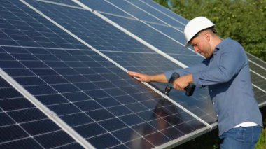 Male engineer in protective helmet installing solar photovoltaic panel system using screwdriver. Electrician mounting blue solar module on roof of modern house