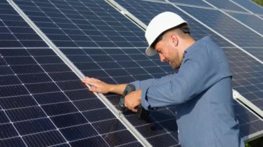 Male engineer in protective helmet installing solar photovoltaic panel system using screwdriver. Electrician mounting blue solar module on roof of modern house. Alternative energy ecological concept.