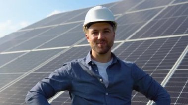 Happy caucasian engineer in helmet smiling at camera at solar power station outside. Positive worker. Ecological industry. Solar farm concept.