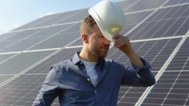 Happy caucasian engineer in helmet smiling at camera at solar power station outside. Positive worker. Ecological industry. Solar farm concept.