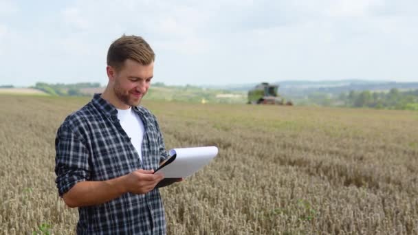 Happy Young Farmer Notebook Standing Wheat Field While Combine Harvester — Vídeo de stock