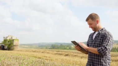 Farmer in wheat field holding digital tablet and showing thumb up. He is smiling. Combine harvester harvesting in background