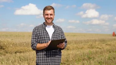 Happy young farmer with notebook standing on wheat field while combine harvester working in background.