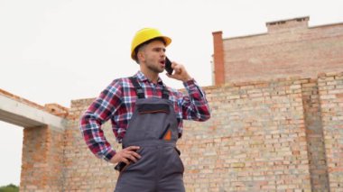 Üniformalı ve güvenlik ekipmanlı inşaat işçisi inşaat işinde. Telefonla konuşuyor..