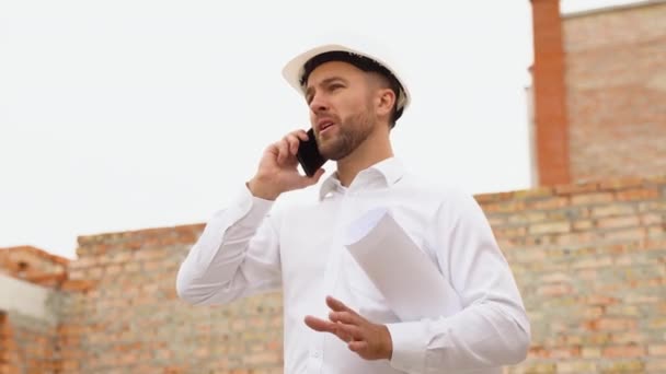 一名戴着白色头盔的土木工程师 手里拿着文件 在电话上谈论着建筑的背景 自然光 — 图库视频影像