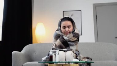 Soğuk algınlığı ve alerji geçiren genç bir kadın şiddetli bir baş ağrısı çekerken battaniyeye sarılı olarak evinde oturuyor. Sağlık hizmetleri kavramı.