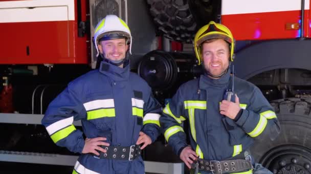 Két Tűzoltó Áll Együtt Egyenruhában Védősisakban Tűzoltóautók Háttérben — Stock videók