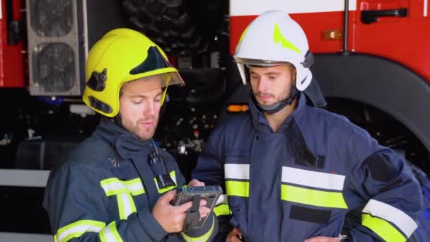 2人の消防士が消防車の外に立ってタブレットを手にし 消火のために何をすべきかを決める — ストック動画