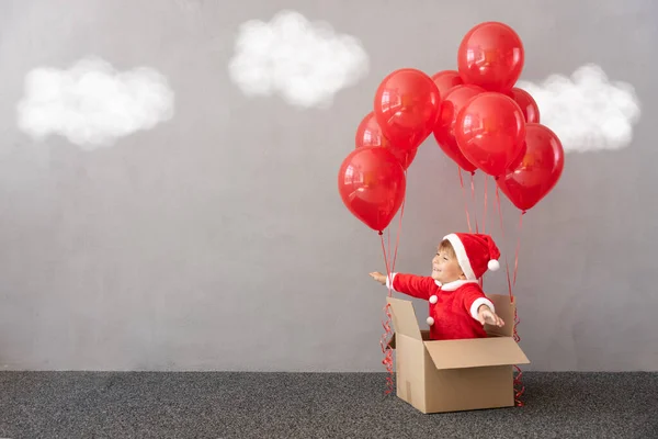 クリスマスの衣装を着た幸せな子供 子供は赤い風船で段ボール箱に座っています 面白い子供が遊んでる 子供が先に飛んでる クリスマスプレゼントボックスのコンセプト — ストック写真