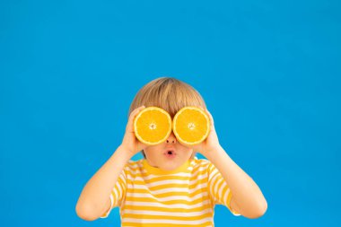 Şaşkın çocuk elinde güneş gözlüğü gibi portakal meyvesi dilimleri tutuyor. Mavi kağıtlı arka planda çizgili sarı tişört giyen bir çocuk. Sağlıklı beslenme ve yaz tatili kavramı