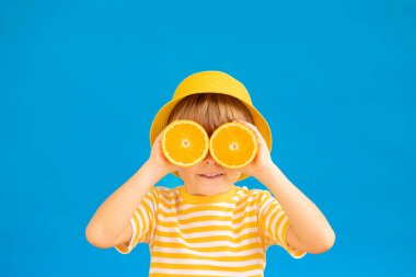 Mutlu çocuk elinde güneş gözlüğü gibi portakal meyvesi dilimleri tutuyor. Mavi kağıtlı arka planda çizgili sarı tişört giyen bir çocuk. Sağlıklı beslenme ve yaz tatili kavramı