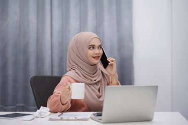 Beautiful asian muslim woman brown hijab drinking coffee and making a phone call while working at her home