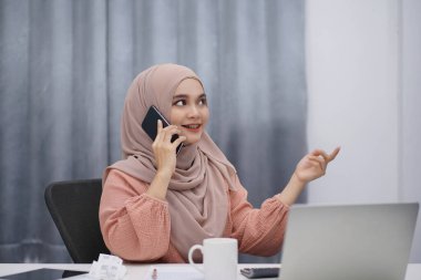 Happy asian muslim woman brown hijab drinking coffee and making a phone call while working at her home