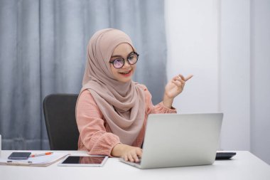 Attractive asian muslim woman in brown hijab having video call with friends or lover, using digital laptop at her home