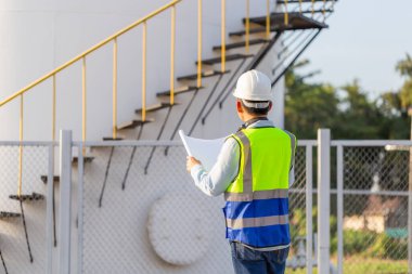 Mühendis denetim altında ve petrol depolama tankını kontrol ediyor, yelek ve kasklı mühendis ve petrol depolama tesisinde belgeleri var.