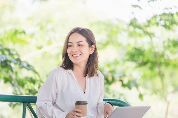 Young freelancer sitting with a laptop in a green park. Smiling modern woman sitting on bench while drinking takeaway coffee