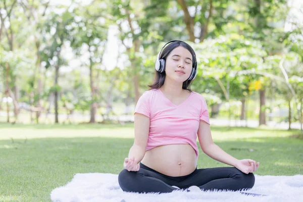 年轻孕妇一边练习瑜伽一边听音乐 怀孕妇女坐在公园里放松 — 图库照片