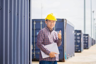 Dizüstü bilgisayarlı gülümseyen mühendis konteynır kargosunda baş parmağını gösteriyor Foreman 'ın kaskı ve güvenlik yeleği kargo kutusundan konteynırları kontrol ediyor.