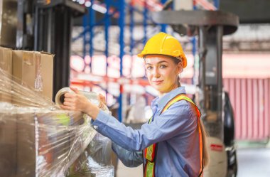 Female worker wrapping boxes in stretch film at warehouse, Worker wrapping stretch film parcel on pallet in factory warehouse