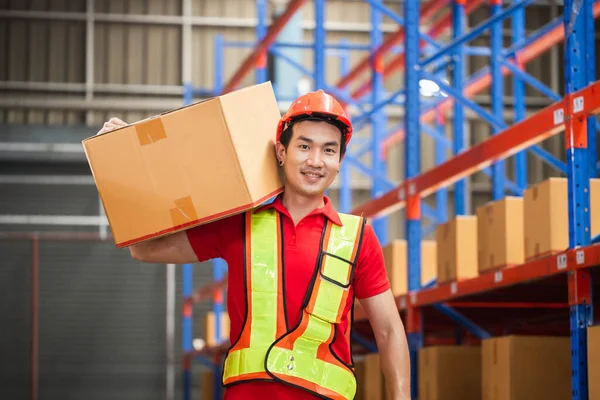 Male worker in hardhat holding cardboard box walking through in retail warehouse, Warehouse worker working in factory warehouse