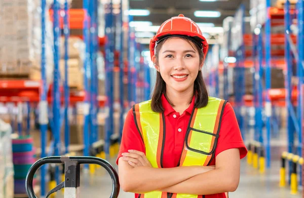 Portrait of young female warehouse worker standing in warehouse with arm crossed, Smiling woman in hard hat looking at camera with arms crossed at warehouse, Worker working in logistic industry inside factory warehouse