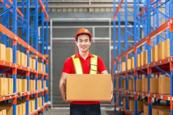 Male worker in hardhat holding cardboard box walking through in retail warehouse, Warehouse worker working in factory warehouse
