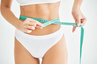 Genç zayıf kadın, beyaz arka planda ölçülü olarak belini ölçüyor.