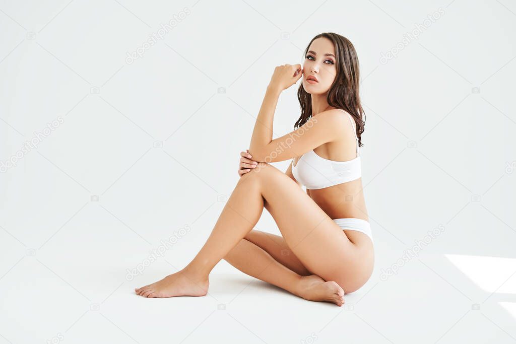 Perfect young woman with sporty body in underwear sitting on floor posing on white studio background