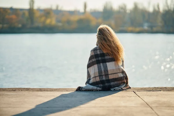 Bakåt syn på ung kvinna avkopplande ensam vid en flod i staden insvept i ull filt njuter av naturen — Stockfoto