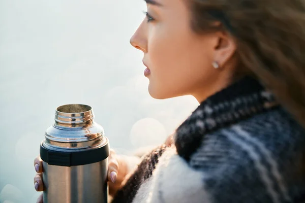 Young pretty woman holds a thermos and drinks tea wrapped in blanket over blur river background. — Stock Photo, Image