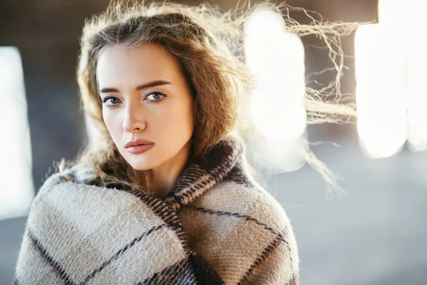 Close-up retrato de bela jovem envolto em cobertor de lã olhando para a câmera no dia ensolarado ventoso no fundo da rua urbana — Fotografia de Stock