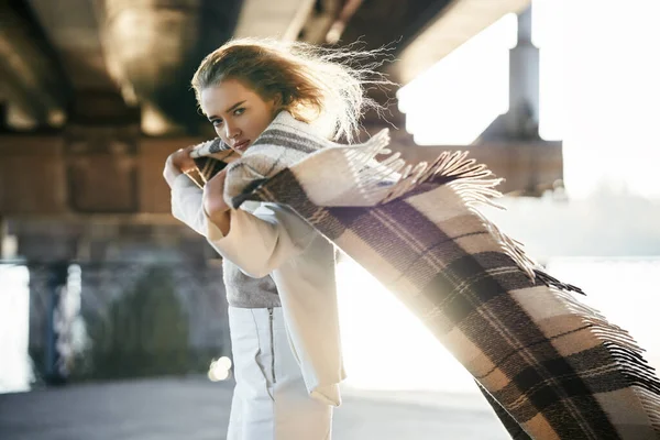 Krásná mladá žena s dekou hledá na kameru na větrné slunečný den pózování na městské ulici pozadí — Stock fotografie