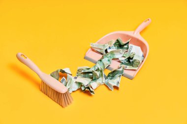 Faraş, el süpürgesi ve buruşuk para dolu sarı arka planlı yaratıcı bir düzen.