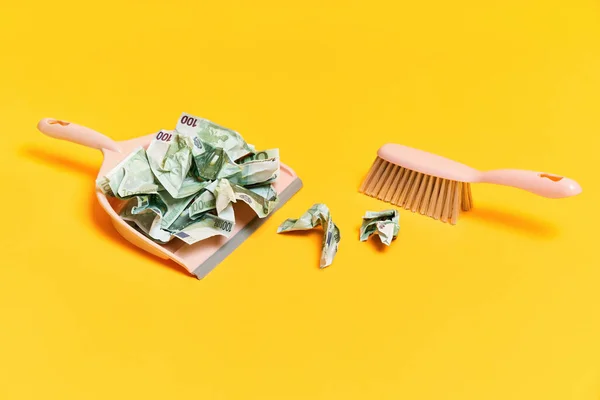 Creative layout with dustpan, hand brooms and crumpled money dollar bills on yellow background — Stock Photo, Image