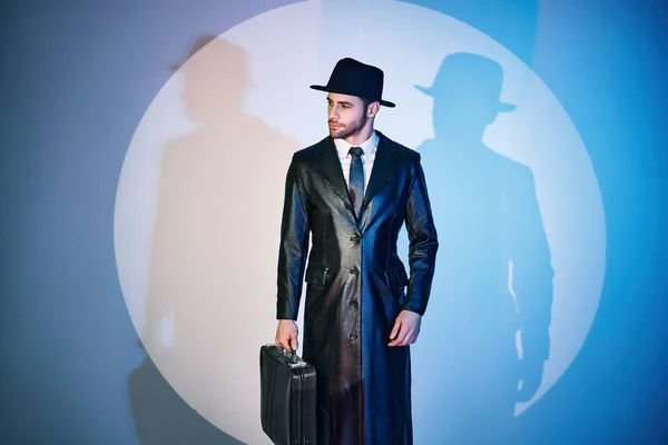 Handsome man in black coat and hat holding briefcase posing in the spotlight on studio background — Fotografia de Stock